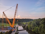 A large yellow crane is positioned near a river, lifting a heavy load. The scene is surrounded by lush green trees and a forest in the background. The sky is partly cloudy with a blend of blue and light pink hues.