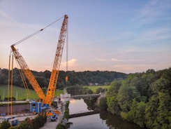 A large yellow crane is positioned near a river, lifting a heavy load. The scene is surrounded by lush green trees and a forest in the background. The sky is partly cloudy with a blend of blue and light pink hues.