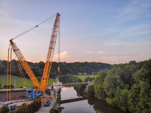 A large yellow crane is positioned near a river, lifting a heavy load. The scene is surrounded by lush green trees and a forest in the background. The sky is partly cloudy with a blend of blue and light pink hues.