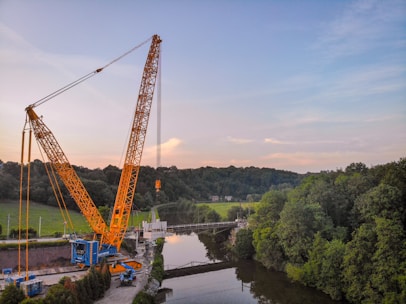 A large yellow crane is positioned near a river, lifting a heavy load. The scene is surrounded by lush green trees and a forest in the background. The sky is partly cloudy with a blend of blue and light pink hues.