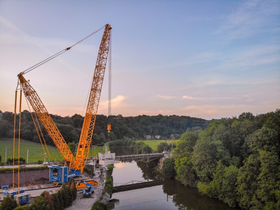 A large yellow crane is positioned near a river, lifting a heavy load. The scene is surrounded by lush green trees and a forest in the background. The sky is partly cloudy with a blend of blue and light pink hues.
