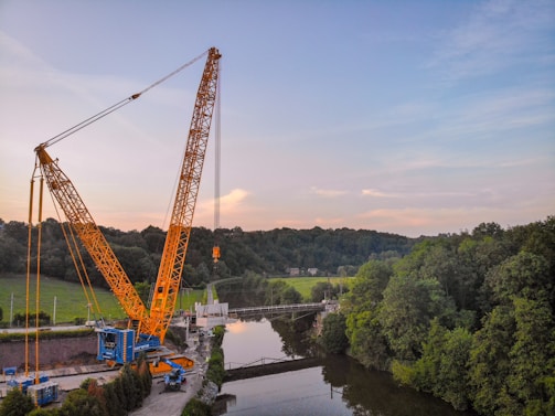 A large yellow crane is positioned near a river, lifting a heavy load. The scene is surrounded by lush green trees and a forest in the background. The sky is partly cloudy with a blend of blue and light pink hues.