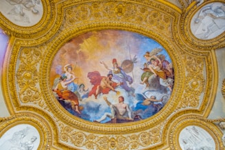 A richly decorated ceiling features a central oval painting surrounded by elaborate gold ornamentation. The painting depicts a group of mythological figures, possibly gods and goddesses, engaged in a dynamic scene with flowing garments and vibrant colors. The gold embellishments around the painting are intricate with floral and foliage motifs.
