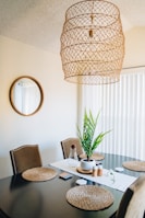 A dining area with a dark wooden table set with woven placemats and a potted plant centerpiece. The walls are neutral-colored, with a large circular mirror adding depth to the space. Above, a unique woven pendant light hangs from the ceiling. Natural light filters in through vertical blinds.