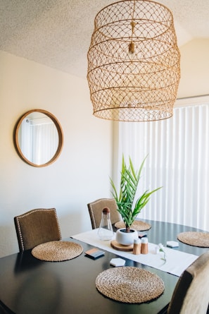 A dining area with a dark wooden table set with woven placemats and a potted plant centerpiece. The walls are neutral-colored, with a large circular mirror adding depth to the space. Above, a unique woven pendant light hangs from the ceiling. Natural light filters in through vertical blinds.