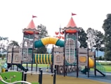 An elaborate playground structure designed to resemble a castle with two towers topped with red cone-shaped roofs and flags. The structure features various climbing elements and slides, including a winding yellow tunnel slide and netted climbing frames. It is surrounded by green and brown ground surfaces, with trees visible in the background.