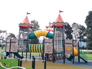 An elaborate playground structure designed to resemble a castle with two towers topped with red cone-shaped roofs and flags. The structure features various climbing elements and slides, including a winding yellow tunnel slide and netted climbing frames. It is surrounded by green and brown ground surfaces, with trees visible in the background.