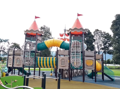 An elaborate playground structure designed to resemble a castle with two towers topped with red cone-shaped roofs and flags. The structure features various climbing elements and slides, including a winding yellow tunnel slide and netted climbing frames. It is surrounded by green and brown ground surfaces, with trees visible in the background.
