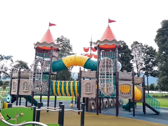 An elaborate playground structure designed to resemble a castle with two towers topped with red cone-shaped roofs and flags. The structure features various climbing elements and slides, including a winding yellow tunnel slide and netted climbing frames. It is surrounded by green and brown ground surfaces, with trees visible in the background.