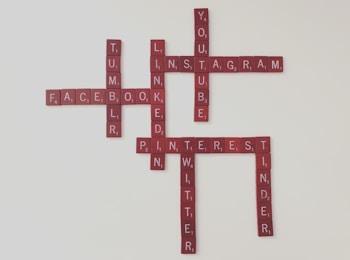 Scrabble tiles spell out names of popular social media platforms, arranged in a crossword style. Names visible include Facebook, Instagram, YouTube, LinkedIn, Pinterest, Tumblr, Twitter, and Tinder. The tiles have a deep red color against a light background.