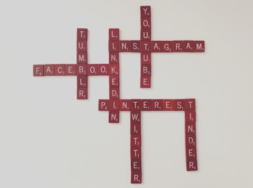 Scrabble tiles spell out names of popular social media platforms, arranged in a crossword style. Names visible include Facebook, Instagram, YouTube, LinkedIn, Pinterest, Tumblr, Twitter, and Tinder. The tiles have a deep red color against a light background.