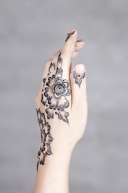 A close-up of a hand with intricate black floral henna designs covering the back of the hand and fingers. The background is blurred and neutral in color.