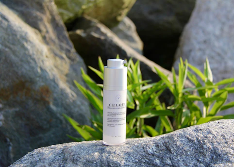 A white cylindrical skincare bottle stands on a large stone, surrounded by greenery. The background is composed of rocks and vibrant green leaves, suggesting a natural setting.