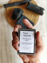 A hand is holding a bar of handmade soap with a label that reads 'Vya Naturals' featuring ingredients like activated charcoal and cedarwood. The background includes pieces of black soap or material placed on a wooden surface.