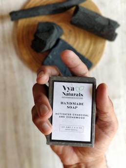 A hand is holding a bar of handmade soap with a label that reads 'Vya Naturals' featuring ingredients like activated charcoal and cedarwood. The background includes pieces of black soap or material placed on a wooden surface.
