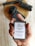 A hand is holding a bar of handmade soap with a label that reads 'Vya Naturals' featuring ingredients like activated charcoal and cedarwood. The background includes pieces of black soap or material placed on a wooden surface.
