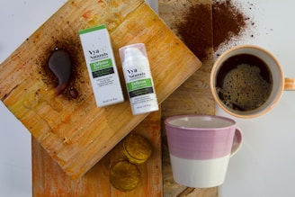 A flat lay composition featuring a box and bottle of caffeine eye cream placed on a textured wooden surface. Next to the cream is a cup filled with coffee, surrounded by coffee grounds and stains. There is also an empty mug with a pink and white design and some small biscuit-like items.