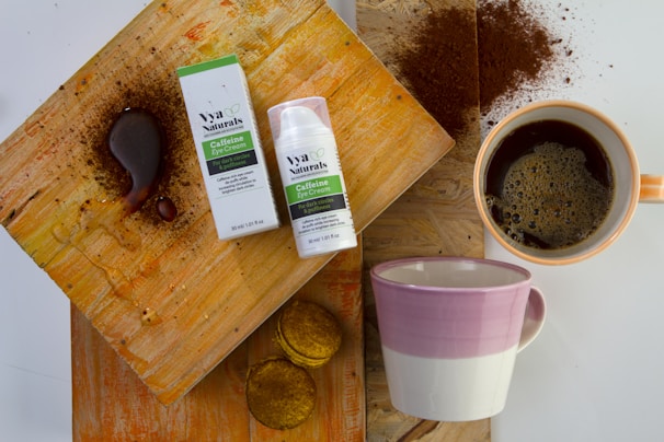 A flat lay composition featuring a box and bottle of caffeine eye cream placed on a textured wooden surface. Next to the cream is a cup filled with coffee, surrounded by coffee grounds and stains. There is also an empty mug with a pink and white design and some small biscuit-like items.