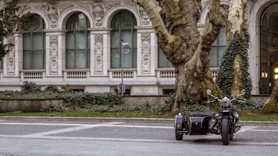 A vintage motorcycle with a sidecar is parked on a cobblestone street in front of a historical building adorned with ornate architectural details and arched windows. Large, mature trees covered in ivy are nearby, contributing to the serene and timeless atmosphere.