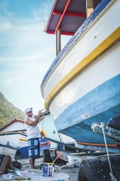 A man is painting the hull of a boat, which is mounted on a trailer. He is wearing casual clothes, including a cap, and is using a brush to apply blue paint. In the background, there are other boats and a hillside. The sky is clear with a few clouds.