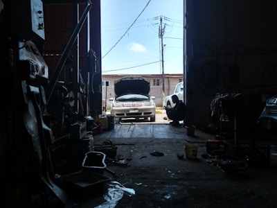 Inside a dimly lit garage, a car is parked with its hood open, suggesting it may be undergoing maintenance or repair. Various tools and auto parts are scattered around the floor. A silhouette of a person is sitting on the right, possibly working or taking a break. Sunlight streaming in from the open door casts bright light on the vehicle.