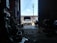Inside a dimly lit garage, a car is parked with its hood open, suggesting it may be undergoing maintenance or repair. Various tools and auto parts are scattered around the floor. A silhouette of a person is sitting on the right, possibly working or taking a break. Sunlight streaming in from the open door casts bright light on the vehicle.
