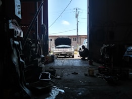 Inside a dimly lit garage, a car is parked with its hood open, suggesting it may be undergoing maintenance or repair. Various tools and auto parts are scattered around the floor. A silhouette of a person is sitting on the right, possibly working or taking a break. Sunlight streaming in from the open door casts bright light on the vehicle.