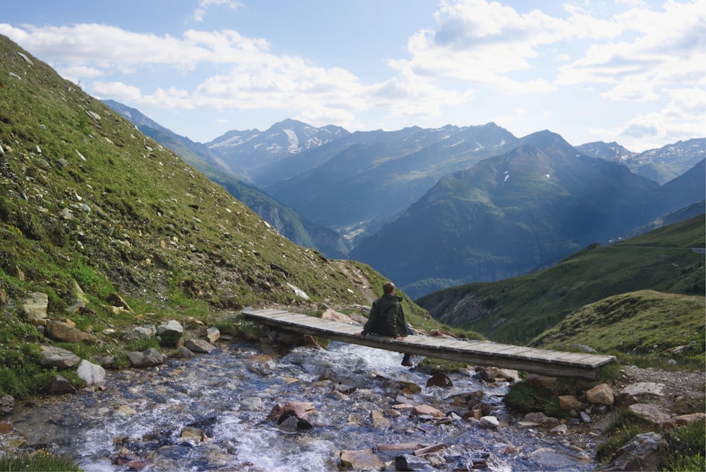 man sitting on plank between waterfalls with view of green mountain ranges