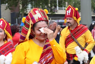 People dressed in traditional colorful garments participate in a cultural parade. The attire features vibrant colors with intricate patterns, and some individuals carry small dolls or similar objects. One woman wipes her face with a tissue, while a man in the background holds a camera, capturing the event.