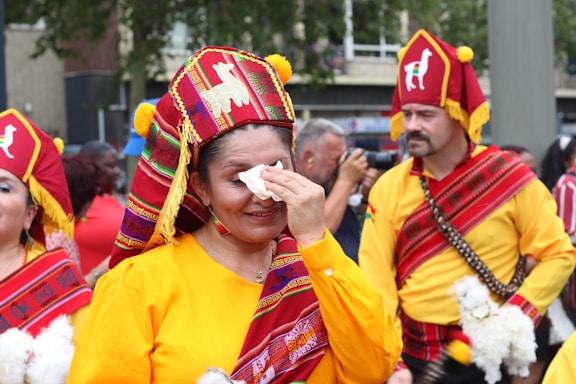People dressed in traditional colorful garments participate in a cultural parade. The attire features vibrant colors with intricate patterns, and some individuals carry small dolls or similar objects. One woman wipes her face with a tissue, while a man in the background holds a camera, capturing the event.