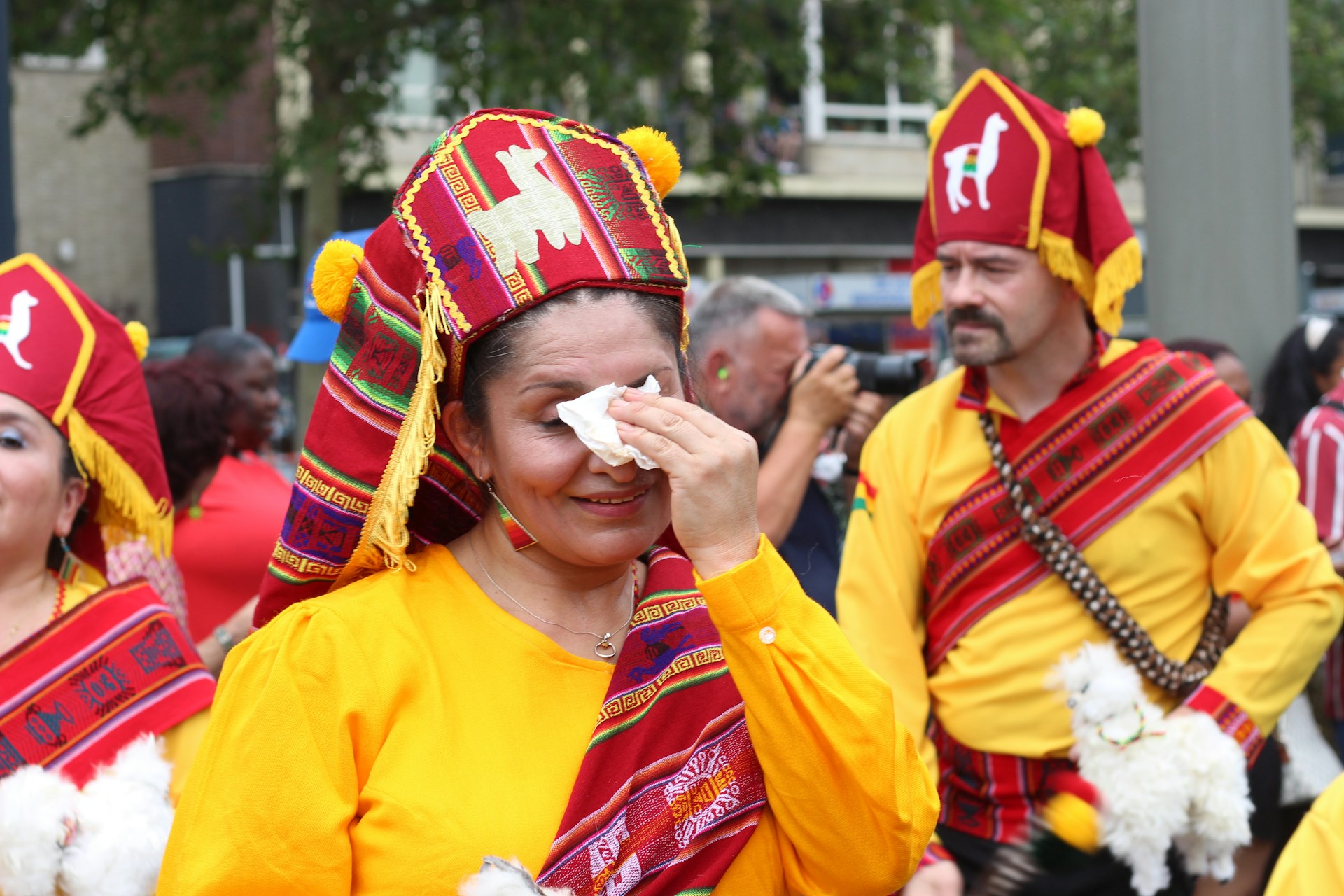 People dressed in traditional colorful garments participate in a cultural parade. The attire features vibrant colors with intricate patterns, and some individuals carry small dolls or similar objects. One woman wipes her face with a tissue, while a man in the background holds a camera, capturing the event.