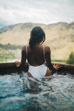A person in a white swimsuit is standing in a round wooden hot tub filled with water. The person is facing away, looking toward a scenic view of mountains and a cloudy sky in the background. The atmosphere is calm and serene, with the pool water gently rippling around the individual.