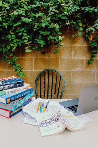 A stack of textbooks is arranged on an outdoor table, surrounded by scattered papers and writing tools, next to an open laptop. In the background, greenery covers a brick wall, adding a natural element to the scene.