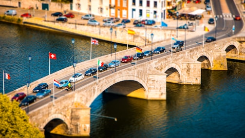 A scenic bridge over a river is seen with various flags from different countries displayed along it. Several cars and a motorbike are crossing the stone bridge, and the surroundings include a riverside with parked cars and buildings.