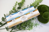 Two boxes of Pharma Hemp Complex natural hemp oil are placed on a surface surrounded by green leafy plants and moss. One box has a blue and white design, while the other features a gold and white design.
