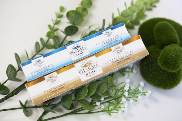 Two boxes of Pharma Hemp Complex natural hemp oil are placed on a surface surrounded by green leafy plants and moss. One box has a blue and white design, while the other features a gold and white design.