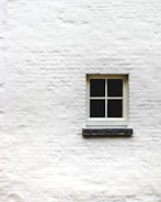 A single window with four panes is positioned in the center of a white brick wall. The wall's texture is rough, and the window frame appears to be made of wood, painted in a light color.