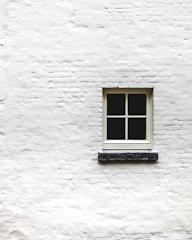 A single window with four panes is positioned in the center of a white brick wall. The wall's texture is rough, and the window frame appears to be made of wood, painted in a light color.