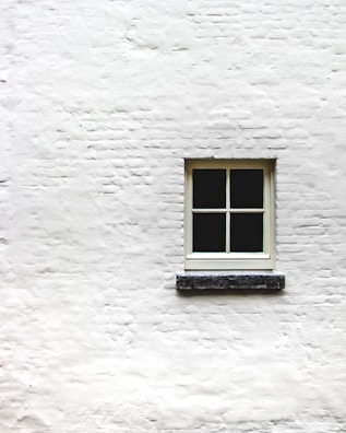 A single window with four panes is positioned in the center of a white brick wall. The wall's texture is rough, and the window frame appears to be made of wood, painted in a light color.