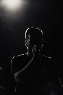A person is silhouetted against a bright overhead light, holding a microphone. The surrounding atmosphere is dark, emphasizing the contrast between light and shadow.