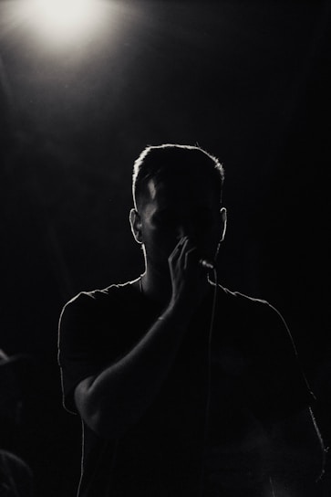 A person is silhouetted against a bright overhead light, holding a microphone. The surrounding atmosphere is dark, emphasizing the contrast between light and shadow.