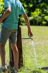 A person wearing a light blue shirt and gray shorts is standing near an outdoor water tap, allowing water to flow onto the grassy ground under bright sunlight. The background is filled with lush green foliage and grass.