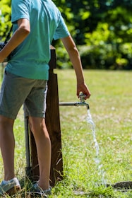 A person wearing a light blue shirt and gray shorts is standing near an outdoor water tap, allowing water to flow onto the grassy ground under bright sunlight. The background is filled with lush green foliage and grass.