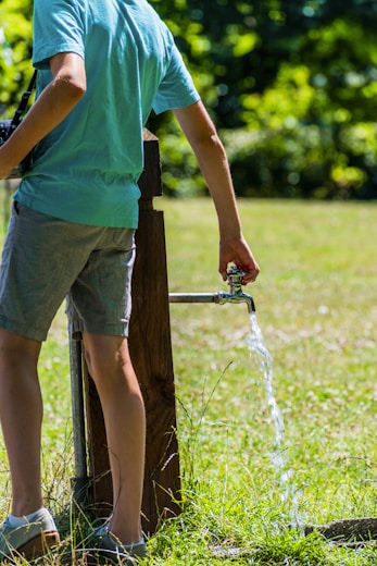 A person wearing a light blue shirt and gray shorts is standing near an outdoor water tap, allowing water to flow onto the grassy ground under bright sunlight. The background is filled with lush green foliage and grass.