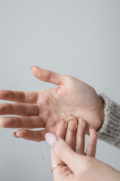 Two hands are depicted, one holding out a delicate gold necklace with a circular pendant, and the other hand is gently holding the necklace. The background is plain and light-colored, creating a minimalist and clean visual.