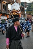 A man dressed in traditional Japanese clothing is holding a long wooden stick. He is wearing a black hat, pink undershirt, and a dark outer robe. Behind him, a group of people dressed in blue and white festival attire are carrying a large ornate structure decorated with white and gold designs. The street is lined with buildings, and there is a festive atmosphere.