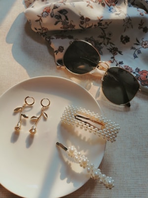 A pair of round sunglasses and a floral-patterned fabric are accompanied by a set of gold and pearl jewelry on a light-colored surface. The jewelry includes a pair of gold dangling earrings shaped like roses and two pearl-embellished hair clips placed on a white plate.