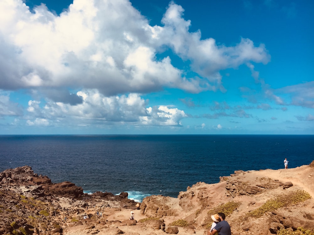 two person near rocky hill viewing blue sea under white and blue skies