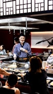 A chef wearing a blue patterned robe and white gloves is cooking in front of a seated audience at a teppanyaki restaurant. The surroundings include a variety of dishes, a grill, and Japanese-style decor with wooden panels and a large calligraphy art piece in the background. Several patrons are engaged, observing the cooking demonstration.