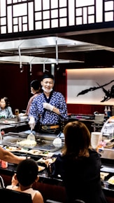 A chef wearing a blue patterned robe and white gloves is cooking in front of a seated audience at a teppanyaki restaurant. The surroundings include a variety of dishes, a grill, and Japanese-style decor with wooden panels and a large calligraphy art piece in the background. Several patrons are engaged, observing the cooking demonstration.